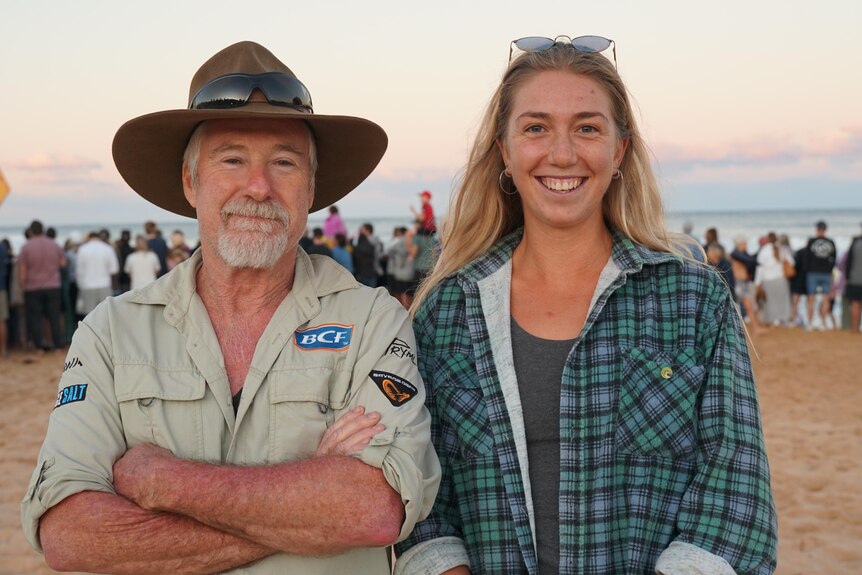 A man wearing khaki and a wide-brimmed hat and a woman wearing a flannel shirt smile at a camera at the beach