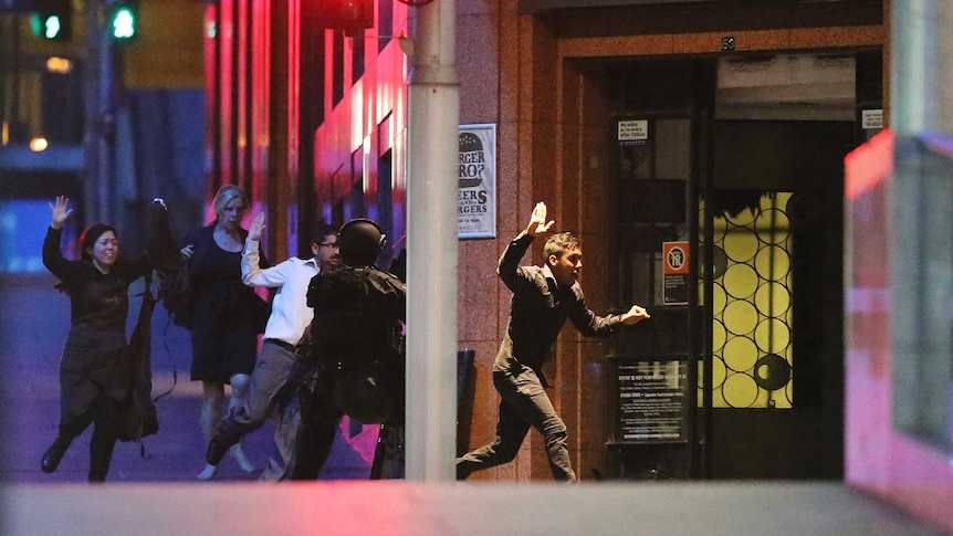 Several people run from the Lindt Cafe with their hands in the air under the watch of a police officer in protective clothing.