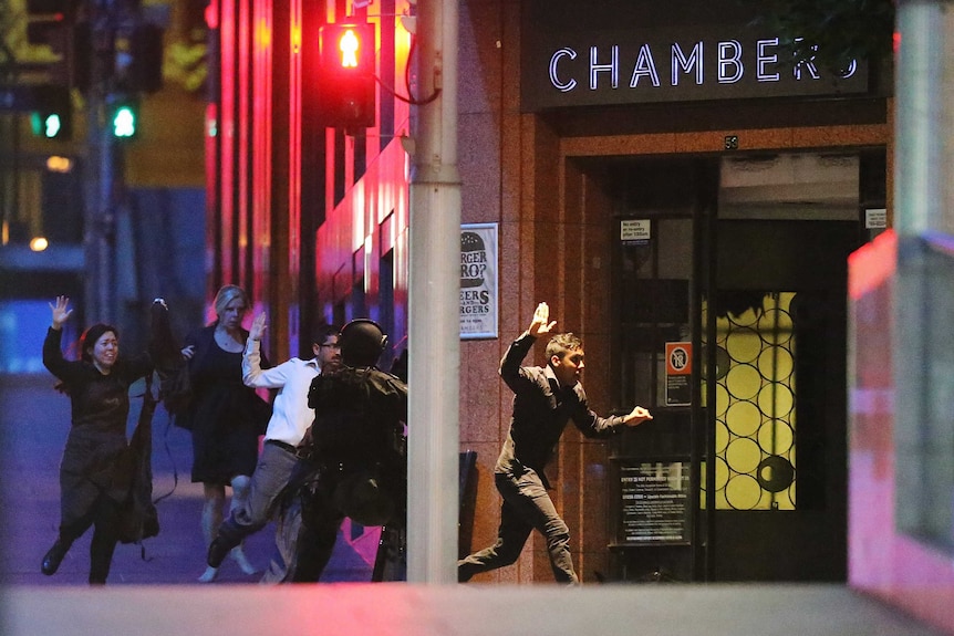 Several people run from the Lindt Cafe with their hands in the air under the watch of a police officer in protective clothing.