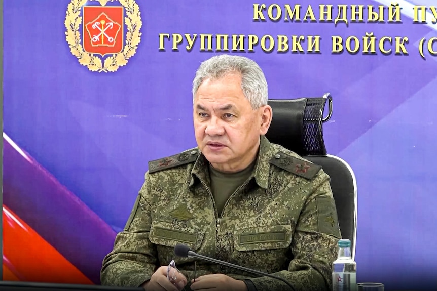 An older man in a military-style jacket without medals sits in front of an official Russian background.
