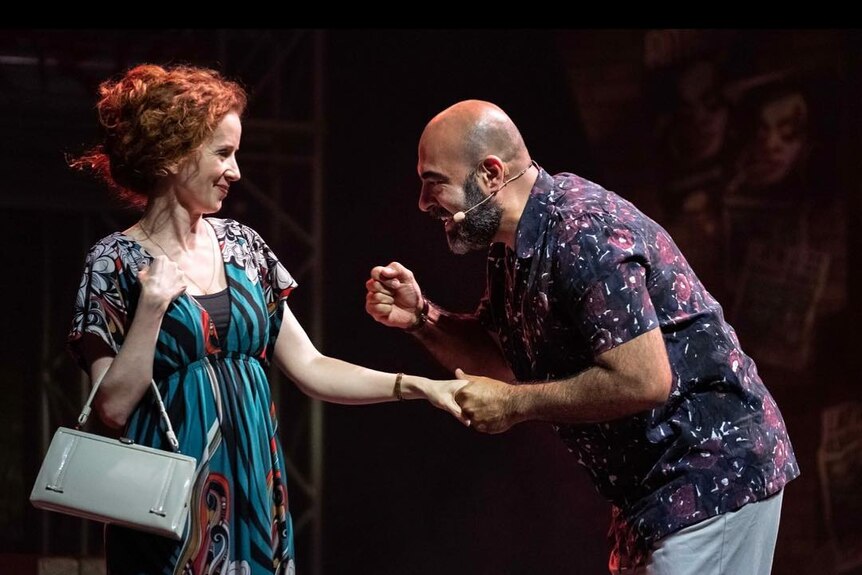 White woman with curly red hair wears a blue patterned dress and clasps hands with Latino man with short beard in dark shirt.
