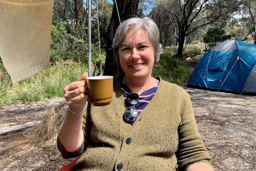 A woman sits on a camping chair holding a mug with a tent in the background.