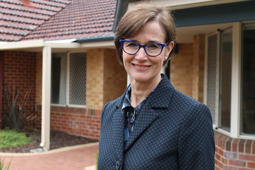 A midshot of a woman smiling for a photo outside a brick house wearing blue spectacles and a blue jacket and shirt.