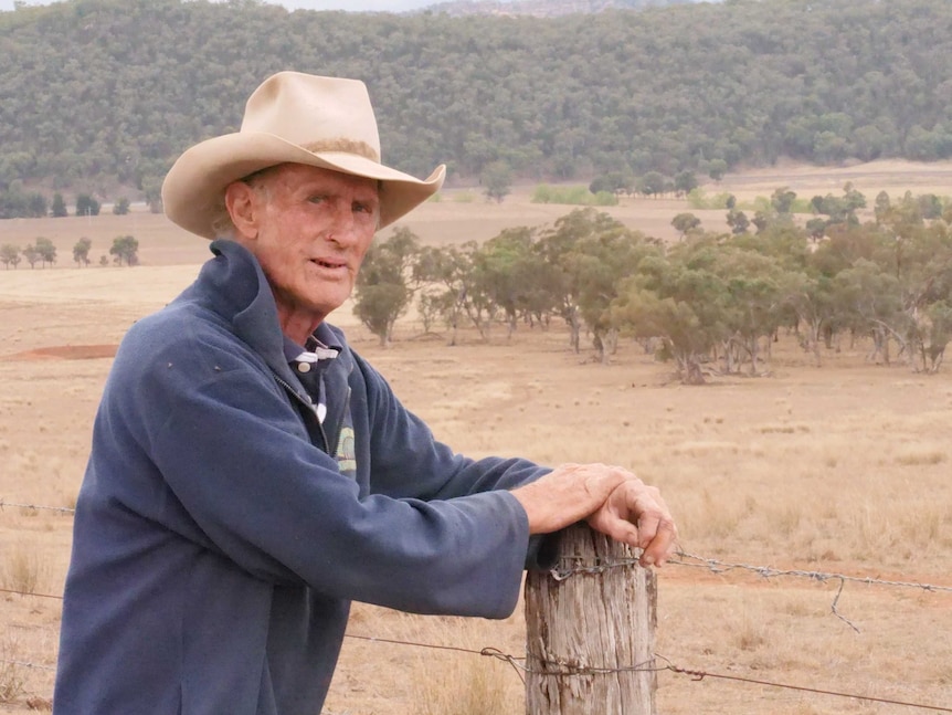 A man in an akubra leans on a farm fence post.