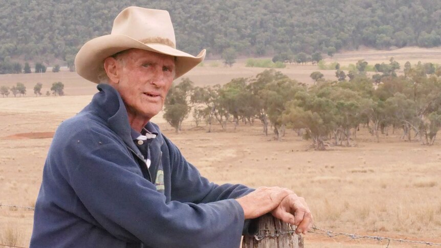 A man in an akubra leans on a farm fence post.