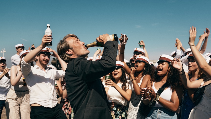 Mads Mikkelsen outdoors surrounded by young people in sailor outfits, throwing head back while chugging on champagne bottle.