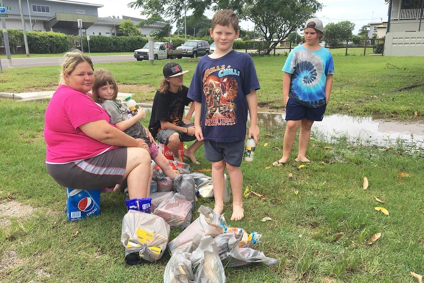 A woman and several children sit and stand on the grass in a street in flooded Ingham.