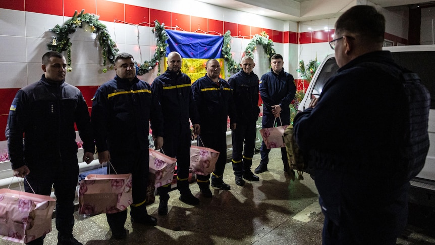 firefighters line up in a fire station holding New Year's gifts as their fire chief speaks to them