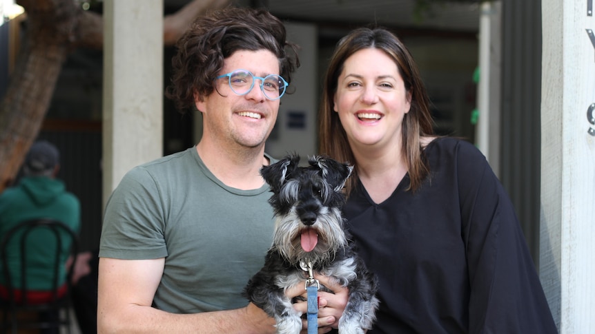 A young dark-haired couple smile at the camera as they sit on a bench, holding their black and grey pet dog
