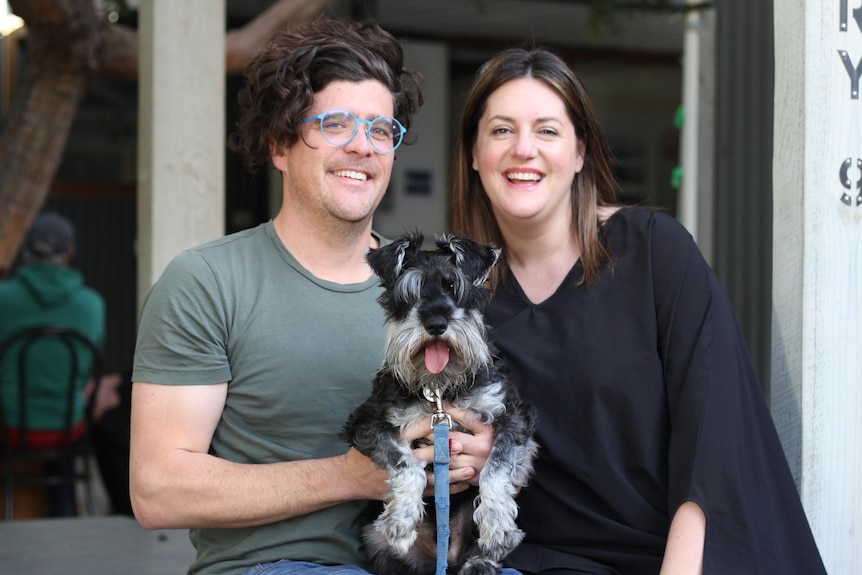 A young dark-haired couple smile at the camera as they sit on a bench, holding their black and grey pet dog