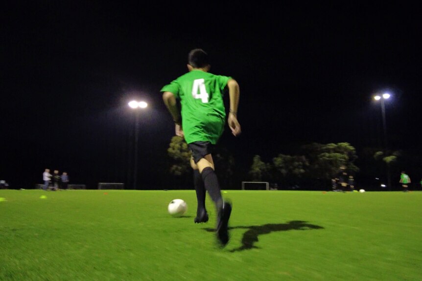 A young man runs away from the camera on a football field at night kicking a soccer ball. He is wearing a team uniform.
