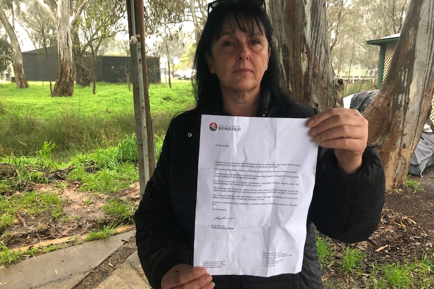 A woman with long, dark hair stands in a park, solemnly holding up an eviction notice.
