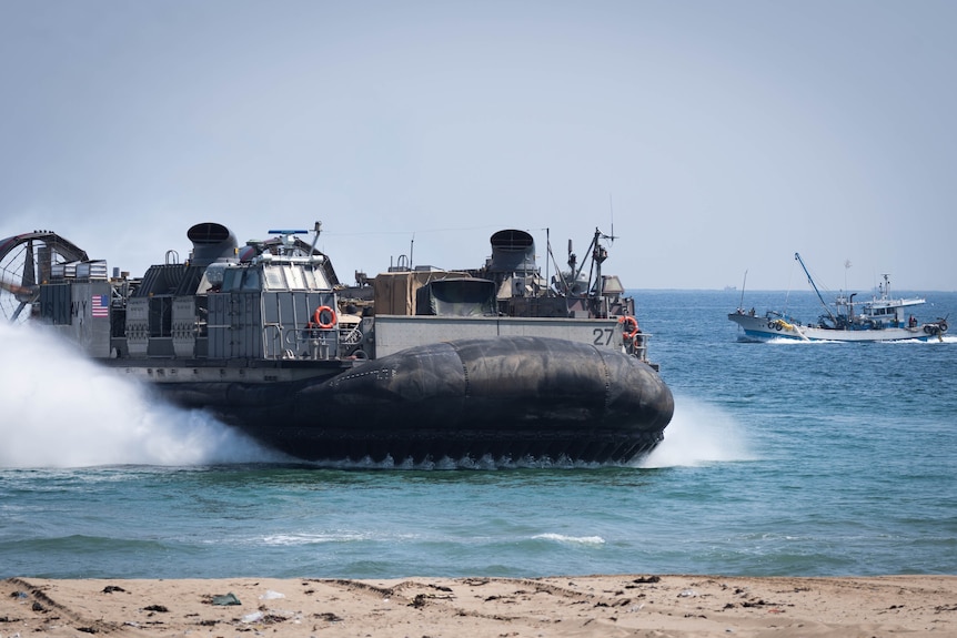 A water-based hovercraft carrying military equipment moves through the ocean water, creating a spray of white 