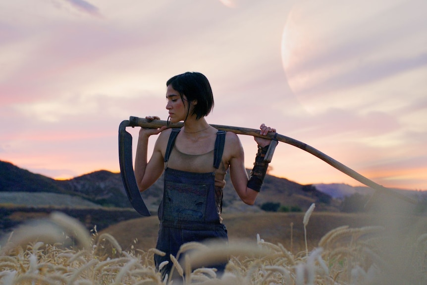 A woman in dirty work clothes holds a scythe while standing in a field.