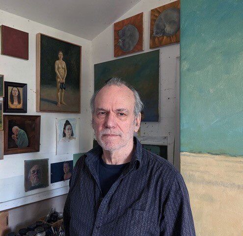 A grey-haired white middle-aged man stands in an art studio wearing a dark navy shirt and surrounded by oil paintings.