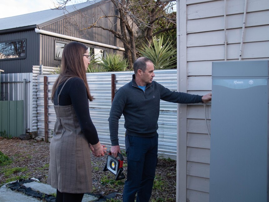 A man touches a pipe outside a house connected to a hot water system, a woman watches