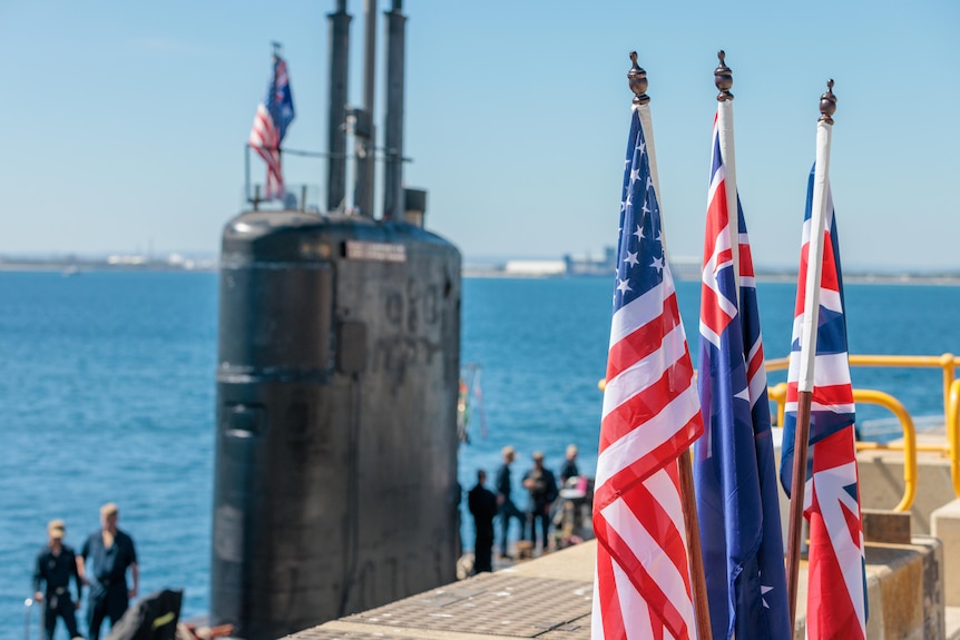 US, Australian and Union Jack flags on poles sit in the foreground, with the top of a submarine at a dock in the background