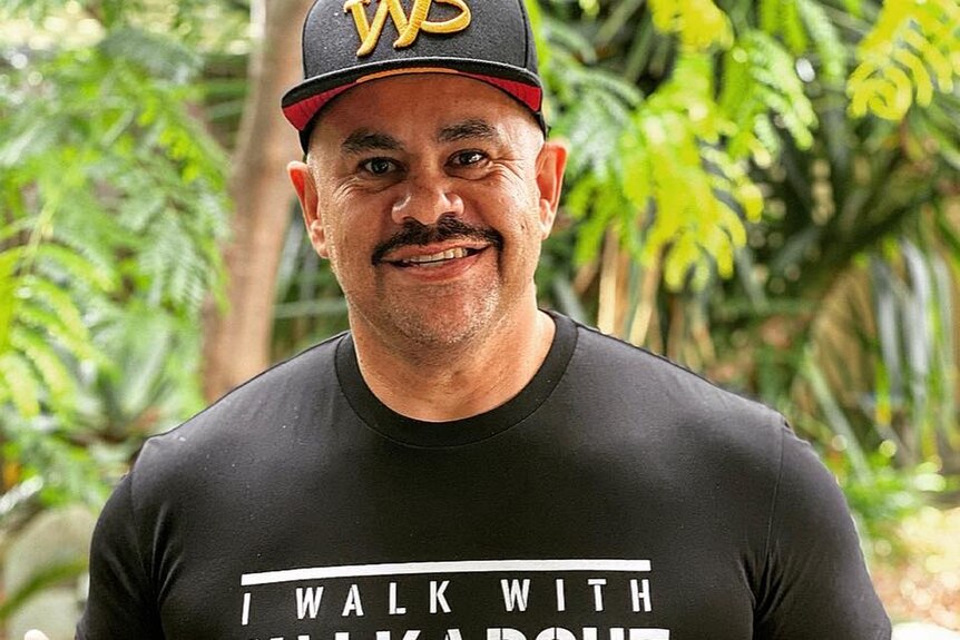 a happy man with a thick brown moustache smiles at the camera with a black shirt and black cap on