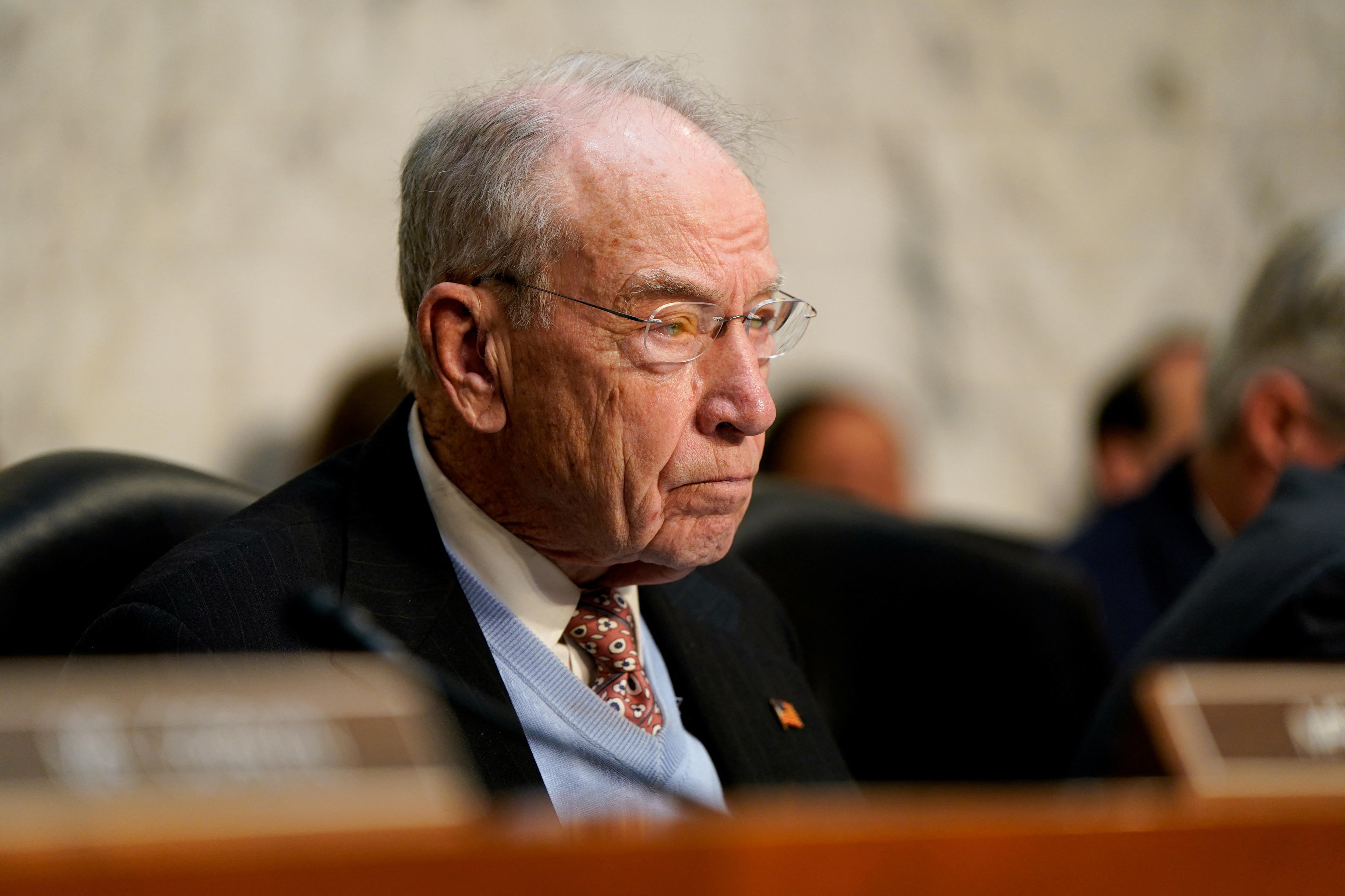 A man in a black suit with a cornflower blue sweater sits with a weary expression