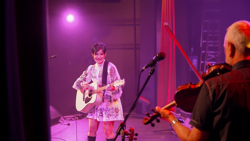 A woman stands on stage under a pink light holding a guitar while a man faces away holding a fiddle