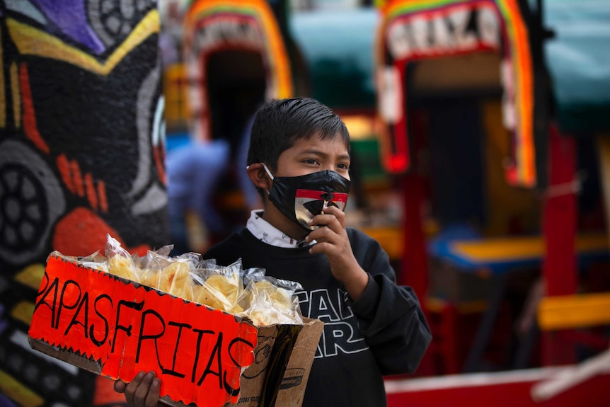 A child, wearing a protective face mask, hawks bags of potato chips near a row of painted wooden boats known as trajineras.