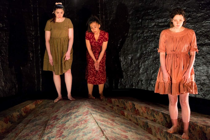 Three women wearing dresses on stage surrounded by darkness.