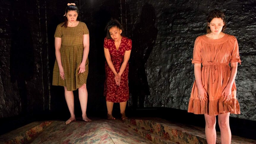 Three women wearing dresses on stage surrounded by darkness.