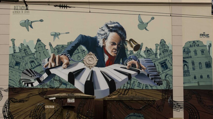 A painted mural on a cream wall. Beethoven is playing a piano with green buildings and flying instruments behind him.