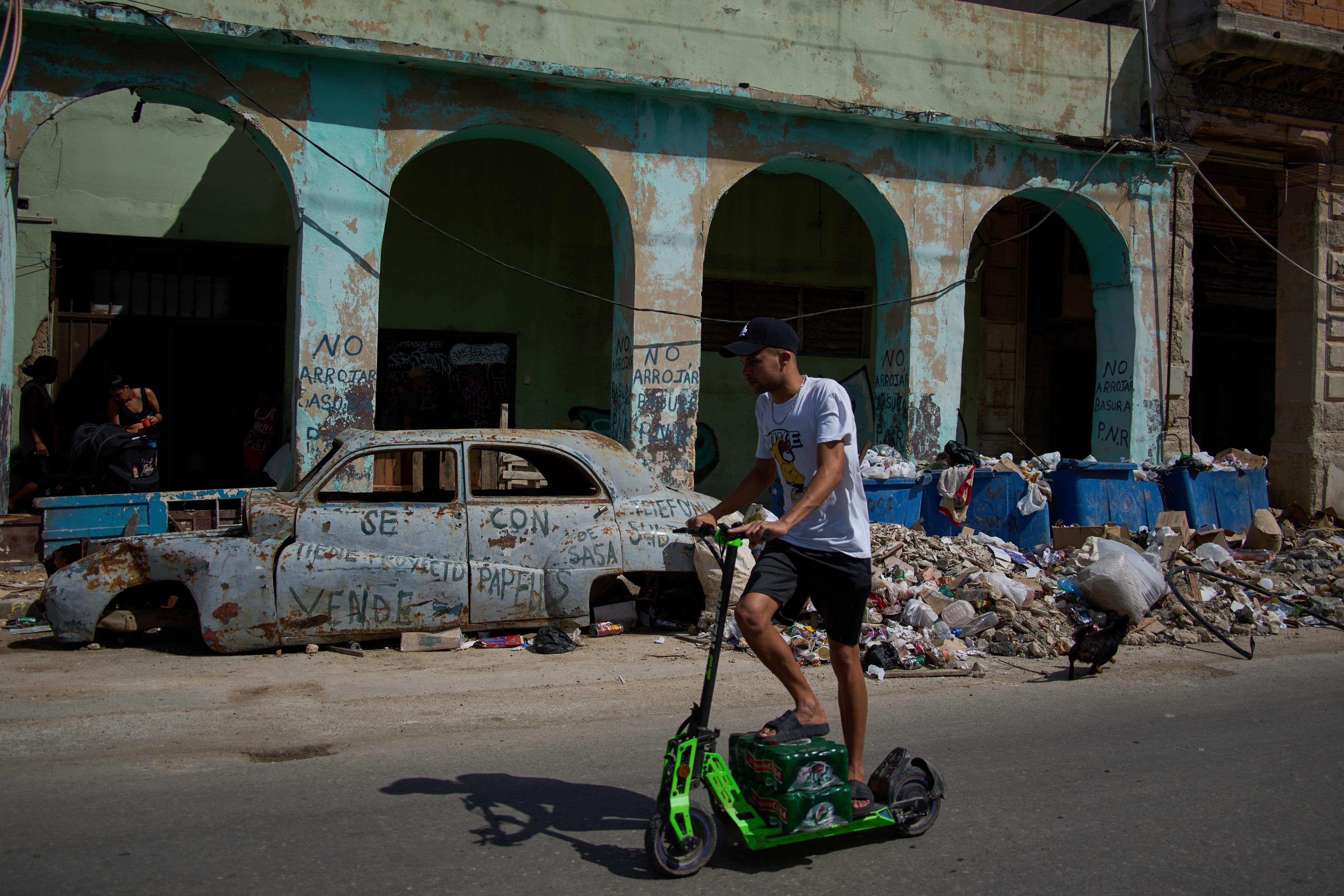 Un cubano conduce una scooter verde por una carretera junto a un coche antiguo oxidado, sin ruedas y con montones de basura.