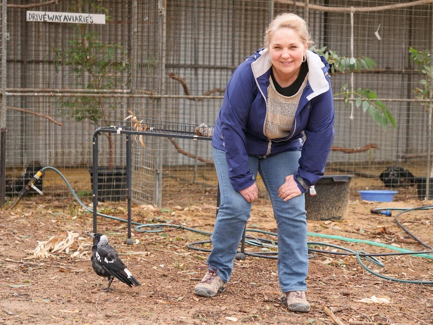 A woman crouching down in a blue jumper and jeans next to a magpie standing in front of bird aviaries.