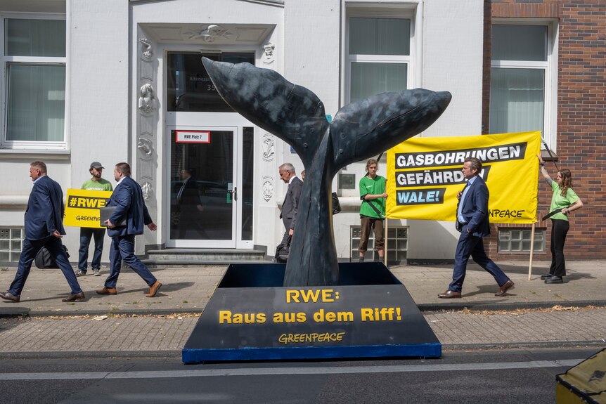 Businessmen walk past activists and a large whale tail sculpture outside RWE's headquarters