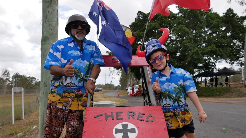 A man and boy pose with their thumbs up next to a billy cart with Aboriginal and Australian flags and a Red Baron theme.