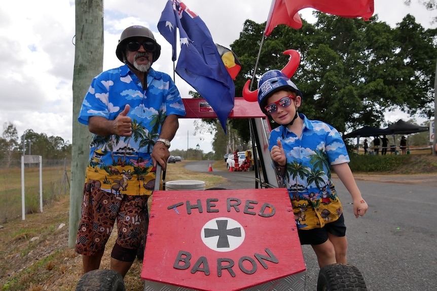 A man and boy pose with their thumbs up next to a billy cart with Aboriginal and Australian flags and a Red Baron theme.