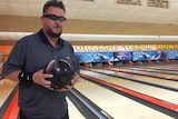 A man with dark glasses holding a bowling ball in front of a tenpin bowling alley
