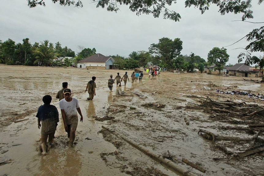 A line of people wade through a muddy field in a village under gloomy skies in a tropical setting.