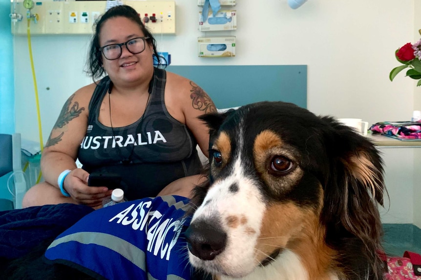 A woman and a cute dog wearing a vest that read 'assistance' sit on a hospital bed.