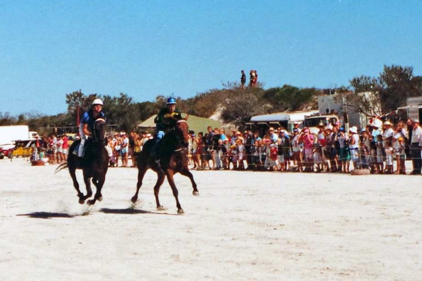 Three horses race along a salt lake, watched by a large crowd.