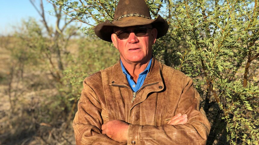 A grazier, wearing a hat and leather jacket, stands in front of a prickly acacia bush