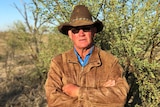 A grazier, wearing a hat and leather jacket, stands in front of a prickly acacia bush