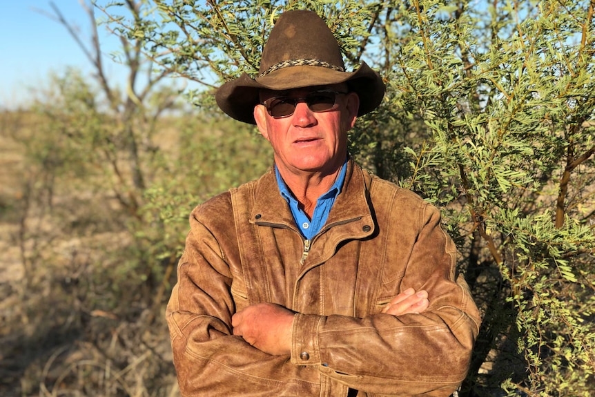 A grazier, wearing a hat and leather jacket, stands in front of a prickly acacia bush