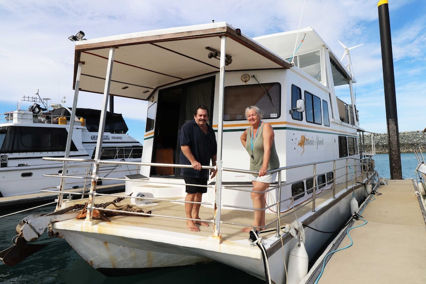 Two people stand on the deck of a boat moored in a marina