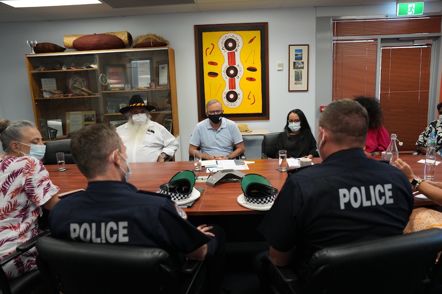 The PM sits at a conference table with two police officers and other people. 