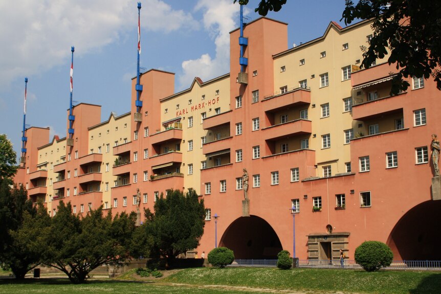 A long, six-storey, brown and yellow apartment building with small turrets and a garden in front