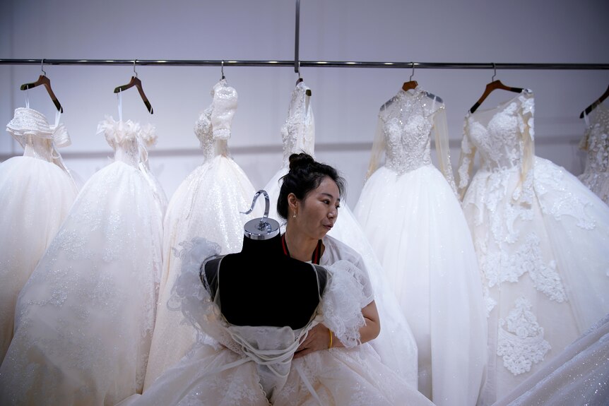 A woman works on a wedding dress on a mannequin with wedding dresses on hangers behind her.