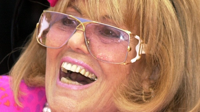Lillian Frank laughs, in a bright pink dress and headpiece with rose-tinted glasses.
