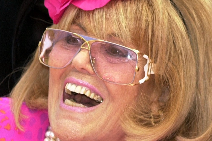 Lillian Frank laughs, in a bright pink dress and headpiece with rose-tinted glasses.