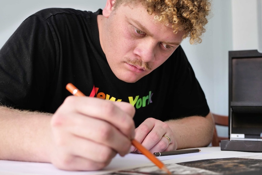 A man sits at a table with a pencil in hand, concentrating on a drawing.