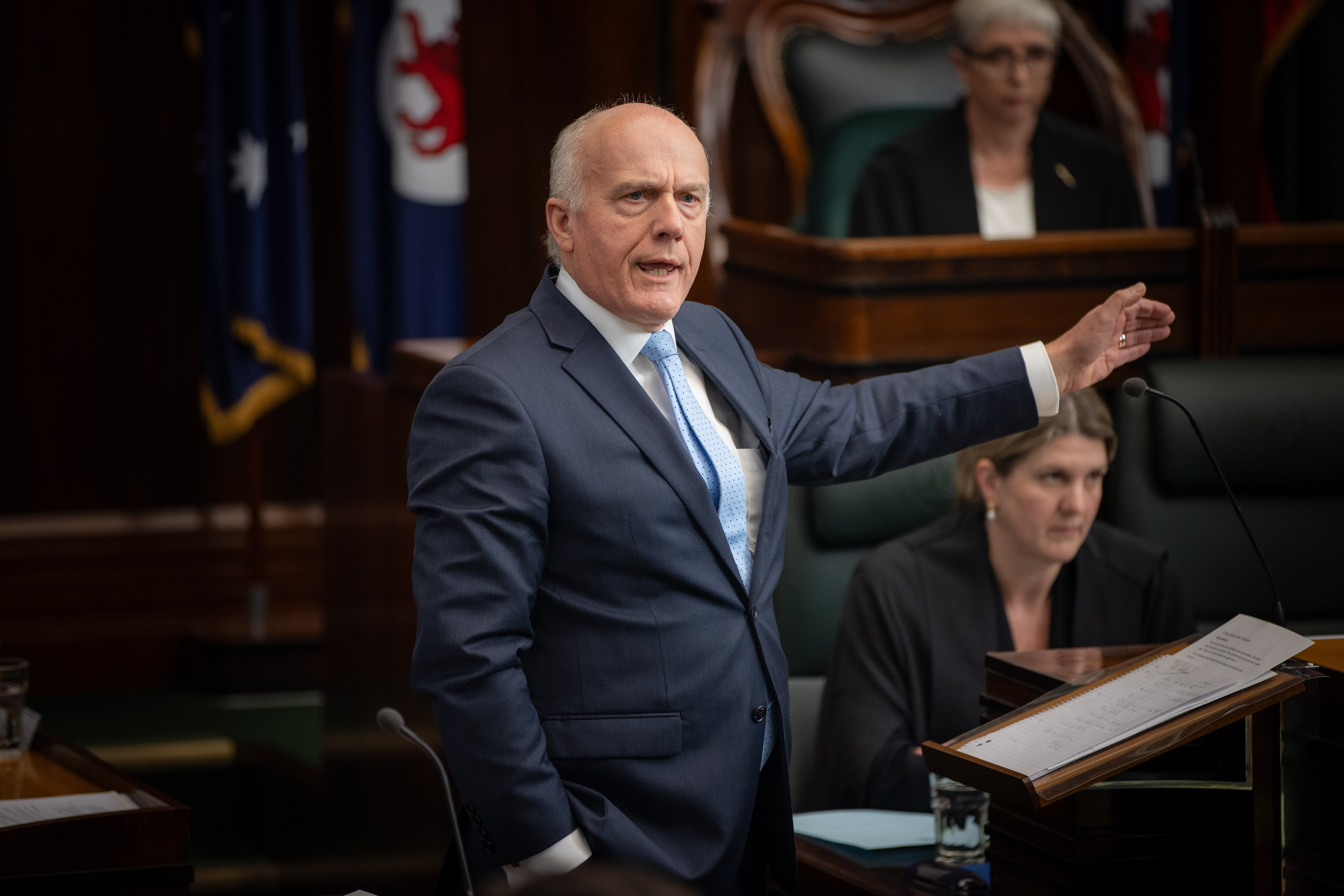 A man in a dark blue suit stands and gestures to his left, addressing a room full of people.