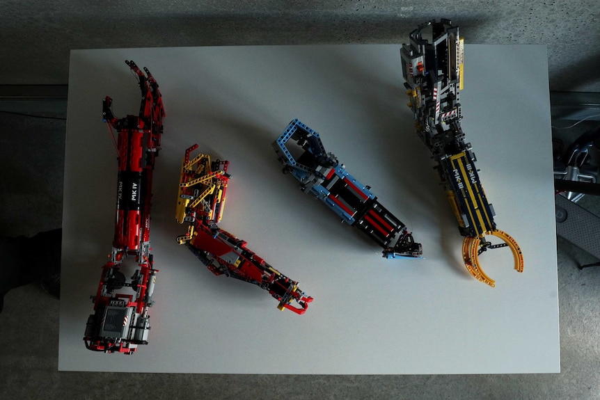 Four different types of prosthetic arms — all made of Lego, all different colours — sit on a table.