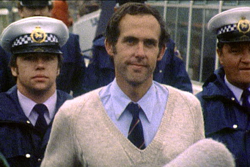 A man in a sweater smiling as he is flanked by police